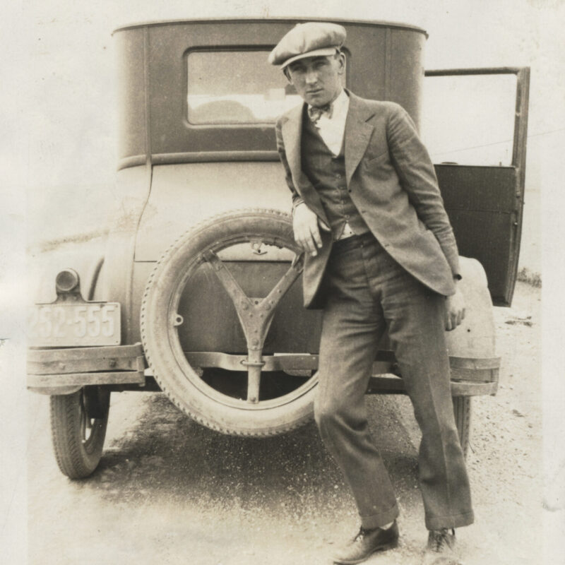 Man in suit leans on vintage car; license plate, handwritten note.