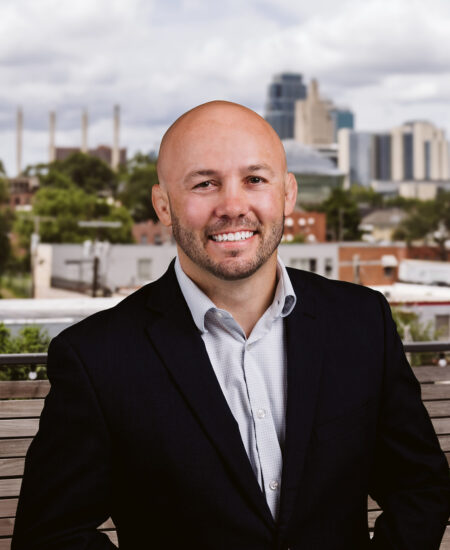 Bald man in suit sits on outdoor bench, city behind.