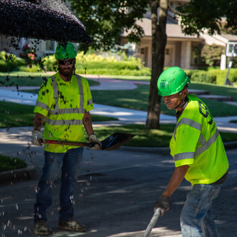 Two workers in yellow vests and green helmets spread asphalt.