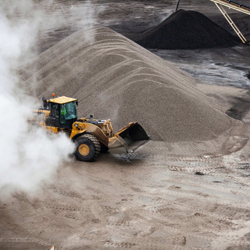 Yellow loader moves gravel by a large pile and rising dust.