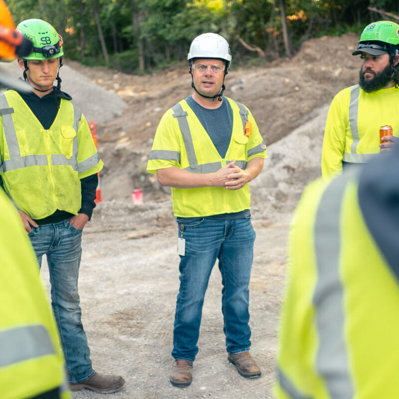 Construction workers in safety gear gather outdoors, one speaking.