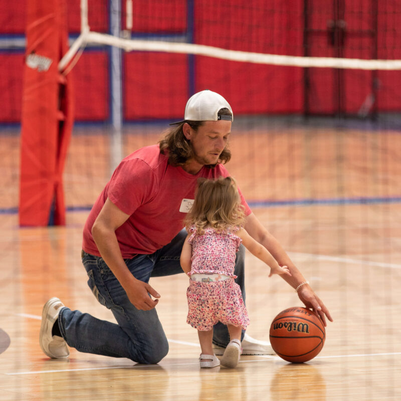 Adult kneels beside child with basketball in gym near nets.