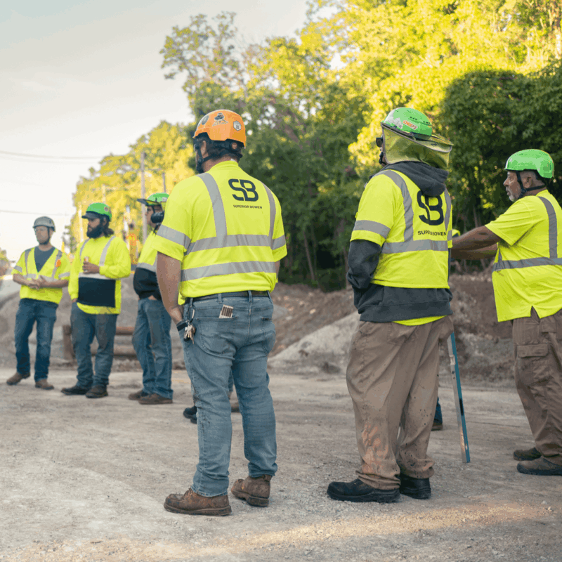 Construction workers in safety gear talk at a tree-lined site.