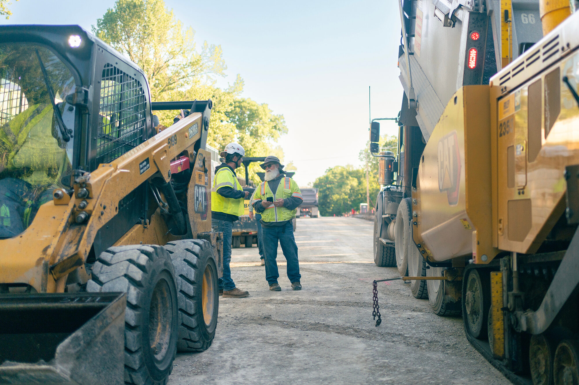 Two construction workers in safety gear stand between heavy machinery.