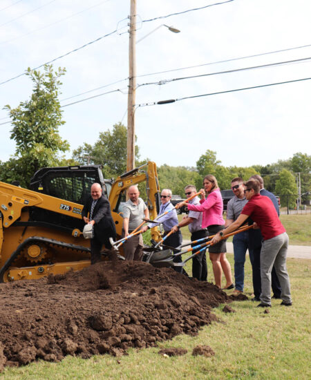 People with shovels at a groundbreaking, equipment and trees behind.