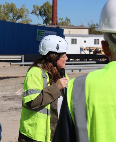 Person in hard hat speaks at industrial site with others nearby.