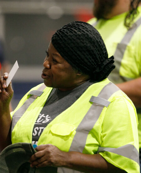 Woman in neon vest with card speaks; another worker behind.