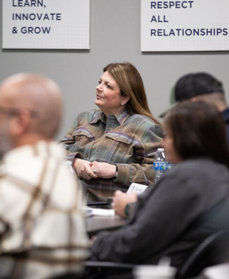 Woman sits in meeting room with others, motivational posters behind.
