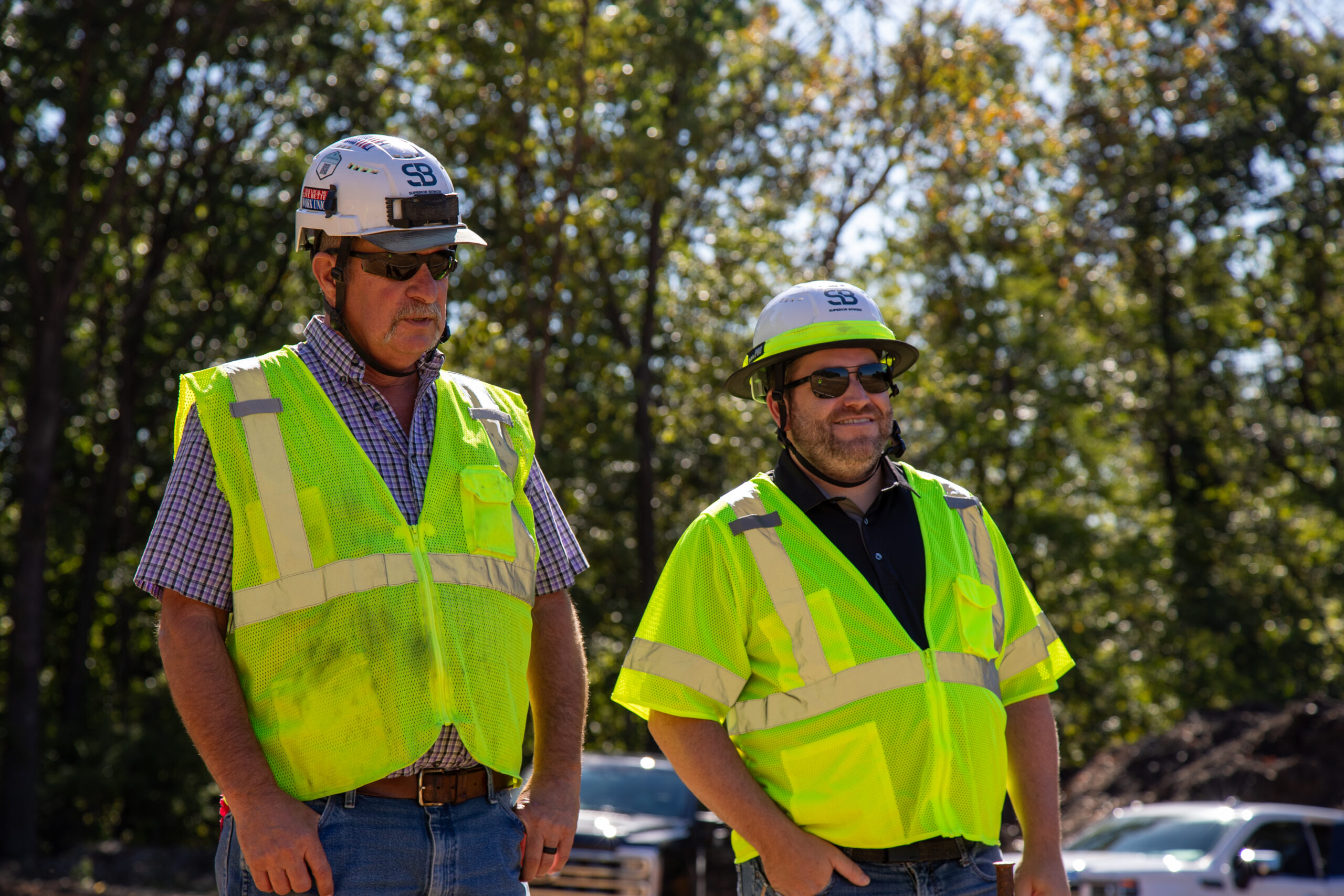 Bearded man in black polo outside, cityscape blurred behind.